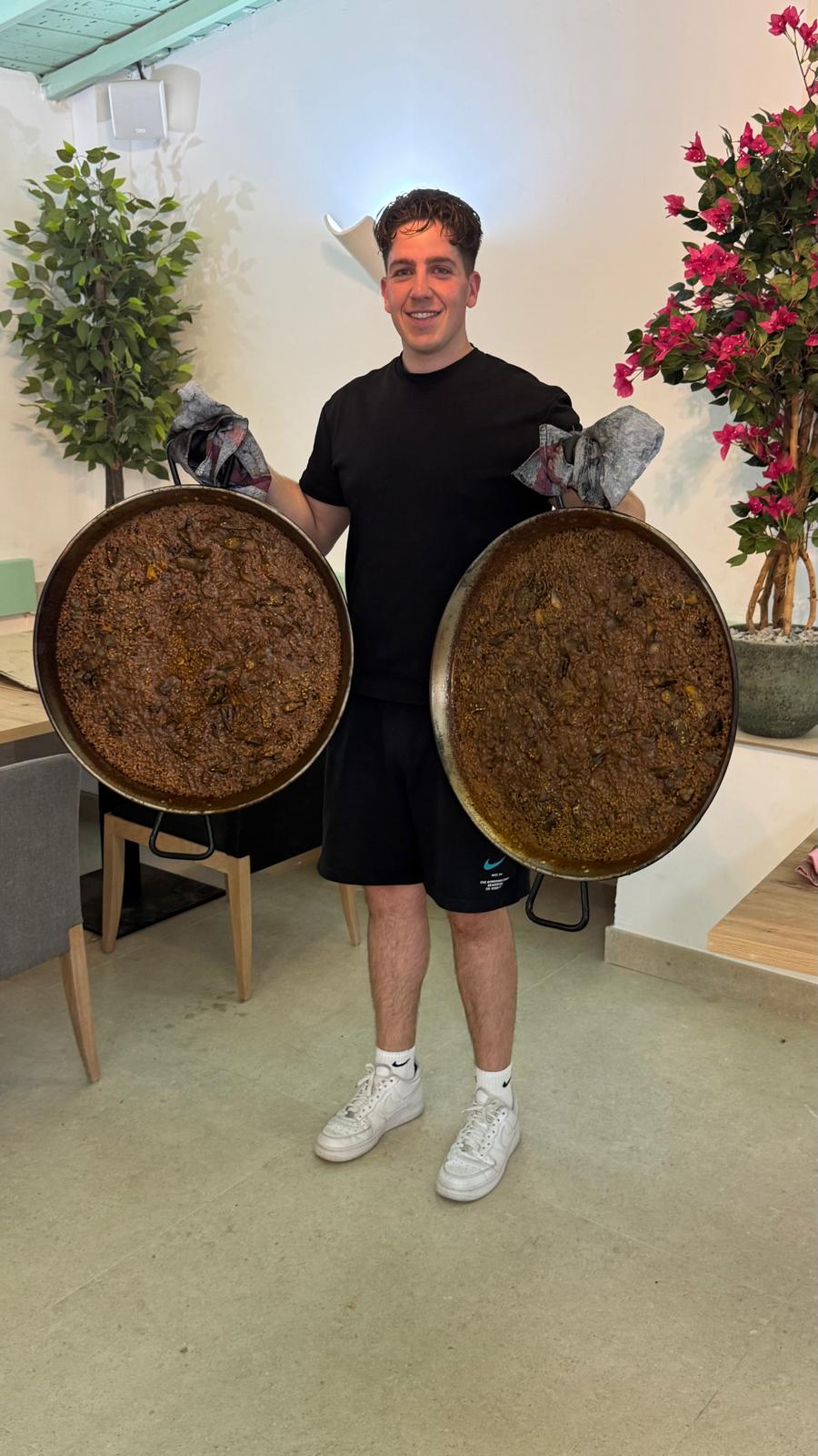Benji, chef del Restaurante Los Pinochos en Simancas (Valladolid), sosteniendo dos grandes paellas recién hechas en la sala del restaurante de cocina valenciana
